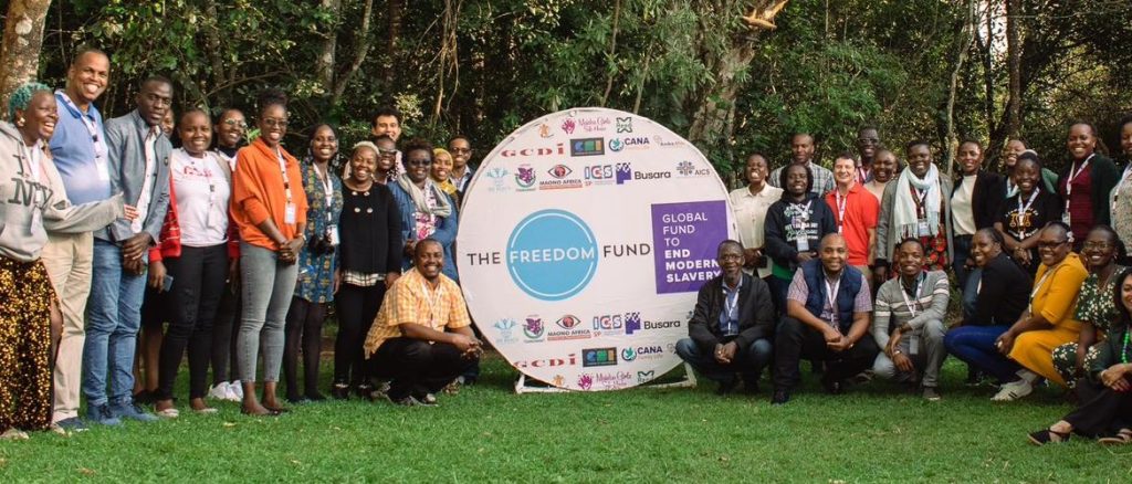 Group photo of a diverse gathering of individuals standing and kneeling on a grassy area in front of a circular sign with the logos of ‘The Freedom Fund’ and ‘Global Fund to End Modern Slavery.’ The backdrop includes lush green trees, and the group members are smiling, appearing engaged and united. Many are wearing lanyards from attending a workshop.