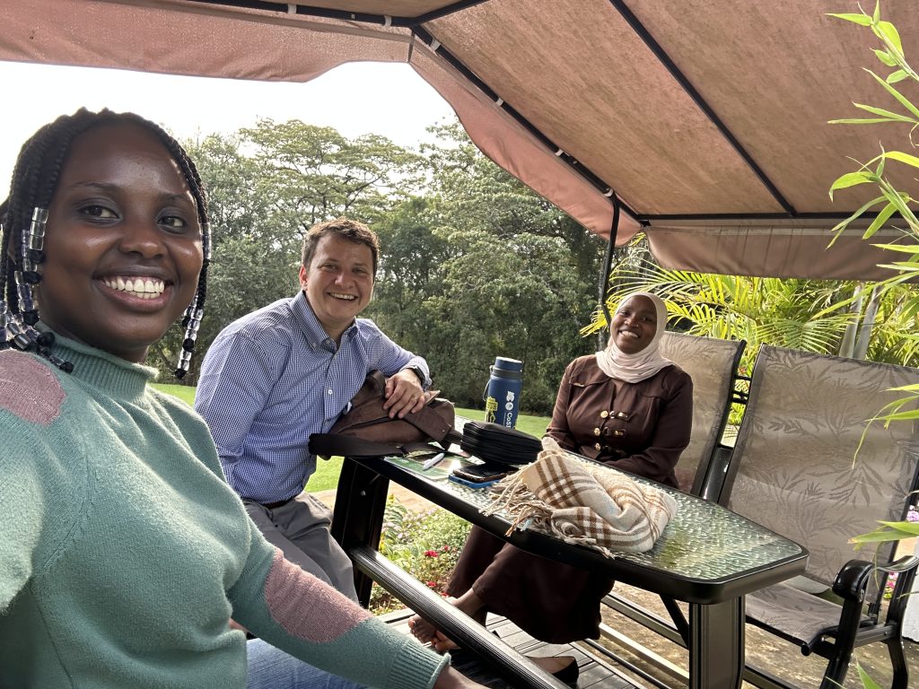 Three individuals sit together at an outdoor table under a covered gazebo, smiling at the camera. The setting is lush and green, with trees and plants in the background. The person on the left wears a green sweater and clear beads in her hair, the person in the middle wears a blue button-down shirt, and the person on the right wears a brown dress with a light hijab/. A thermos, bag, and folded scarf are on the glass table, adding to the relaxed and friendly atmosphere.