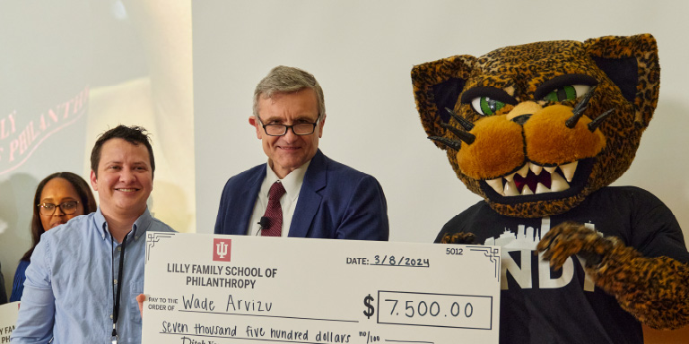 Four individuals pose with a large ceremonial check made out to ‘Wade Arvizu’ for $7,500 from the ‘Lilly Family School of Philanthropy,’ dated March 8, 2024. The group includes a person in a suit, a smiling individual in casual attire, and a mascot wearing a jaguar costume with sharp teeth and intense eyes. The event appears celebratory, with a professional yet lively atmosphere.